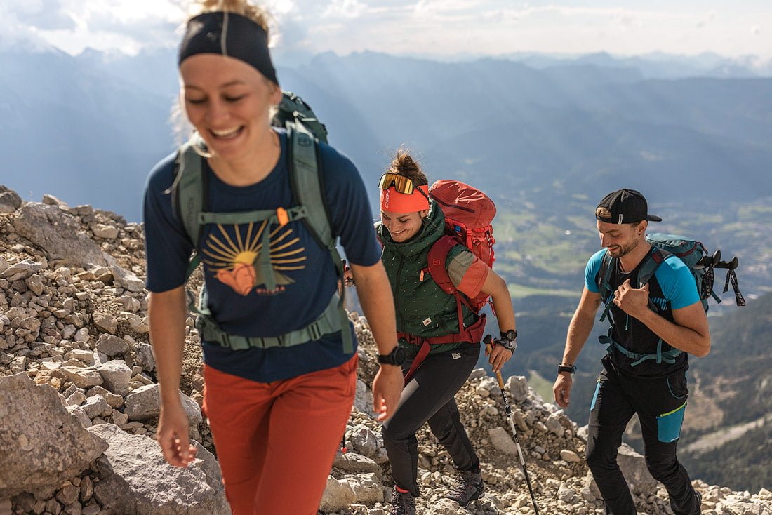 A group of hikers climbing a rocky trail in the Berchtesgaden Alps, smiling and enjoying the challenge with backpacks and hiking gear, against a backdrop of mountain scenery.